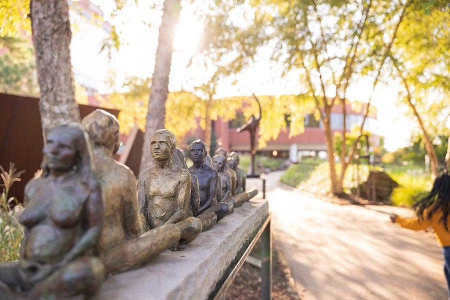 Bronze statue lineup on a sunlit campus path lined with trees.