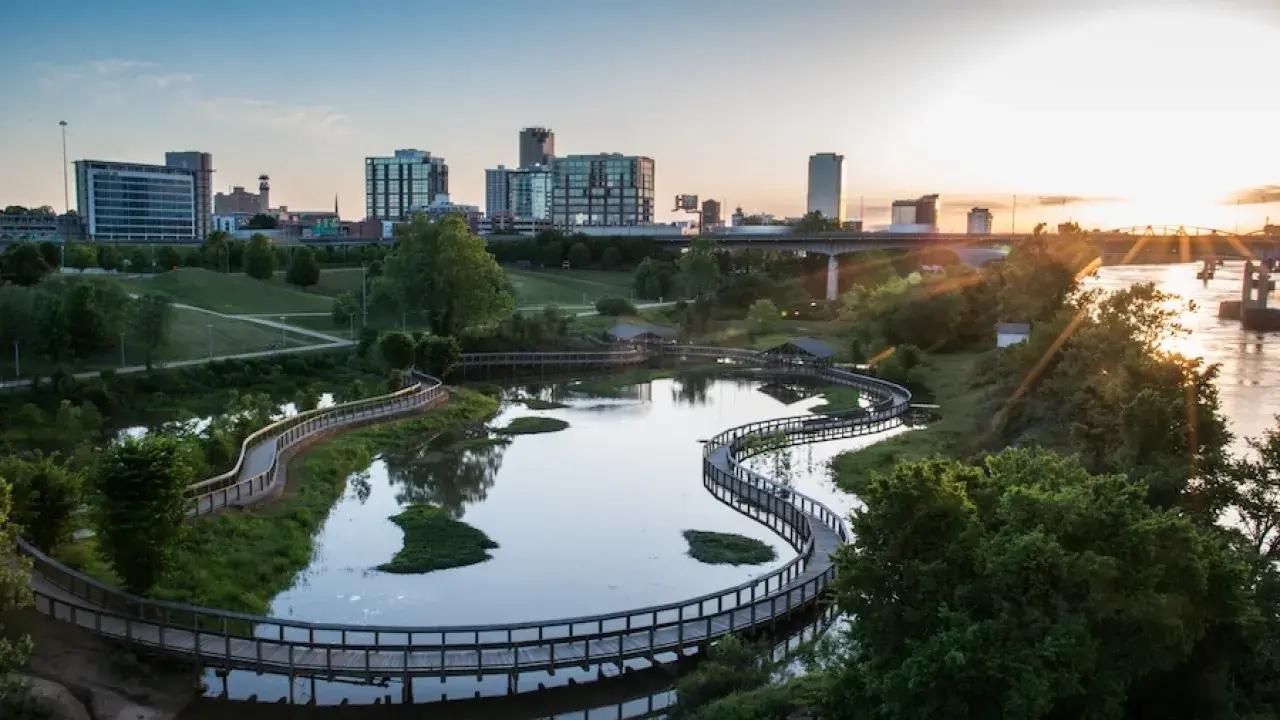 Urban park with winding paths, water features, and city skyline at sunset.