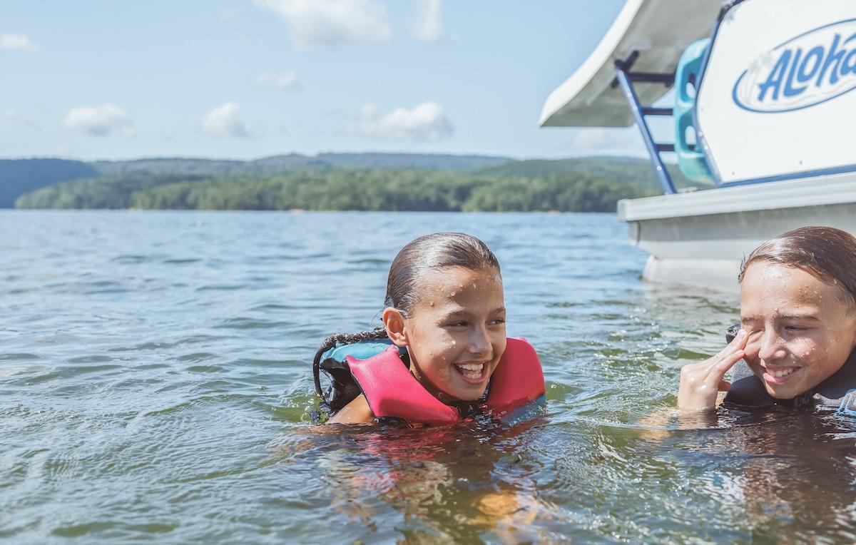 Two kids swimming near a boat, wearing life jackets, smiling on a sunny day.