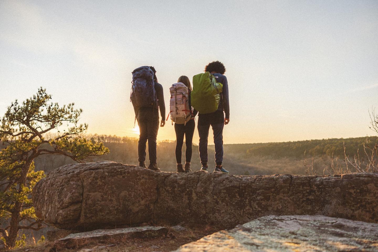 Three hikers with backpacks stand on a rock at sunset.