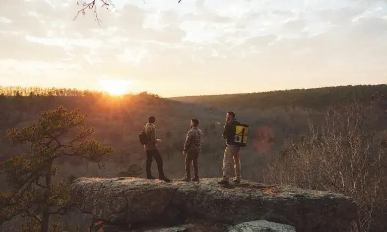 Three hikers stand on a cliff, watching a sunset over a forested landscape.