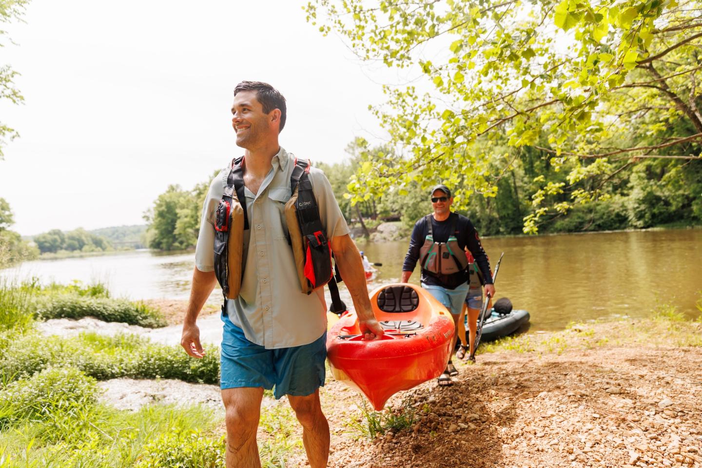 Two men carrying kayaks near a river on a sunny day.
