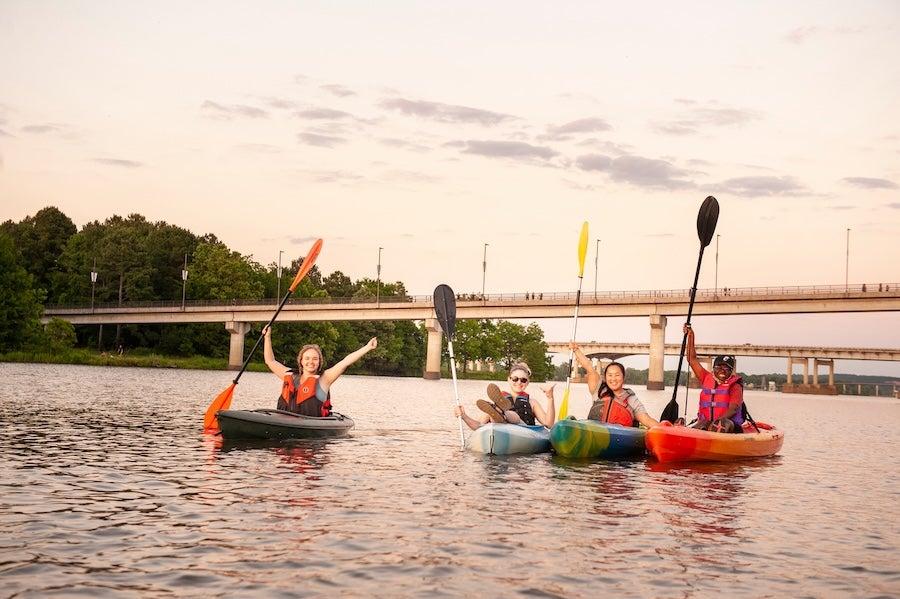 Four people kayaking on a calm river at sunset, bridge in background.
