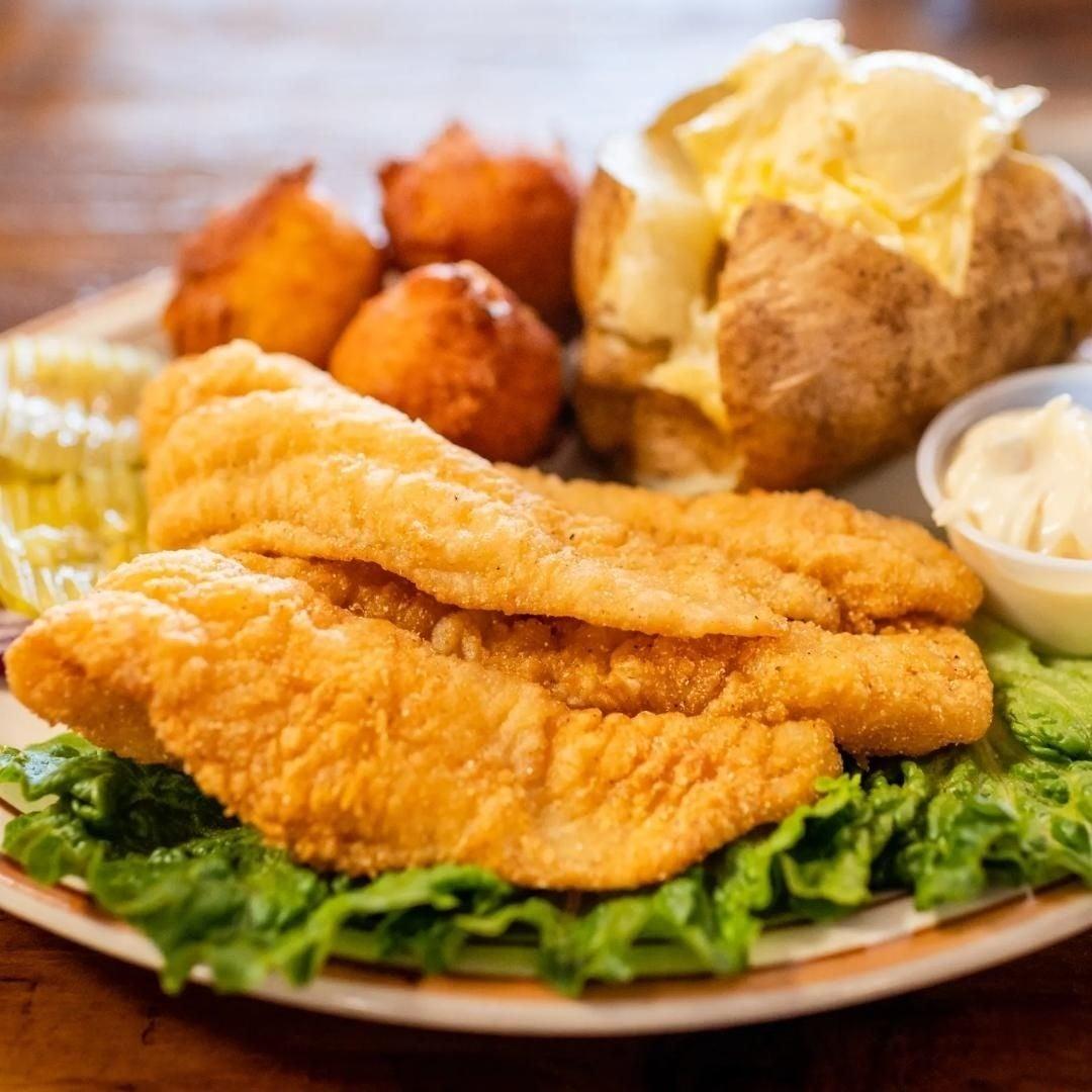 Fried fish with lettuce, baked potato, hush puppies, pickles, and a small sauce cup.