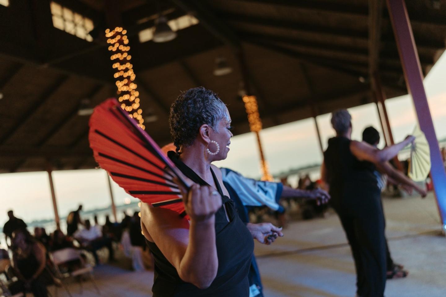 Woman holding a red fan, dancing in a pavilion at sunset.