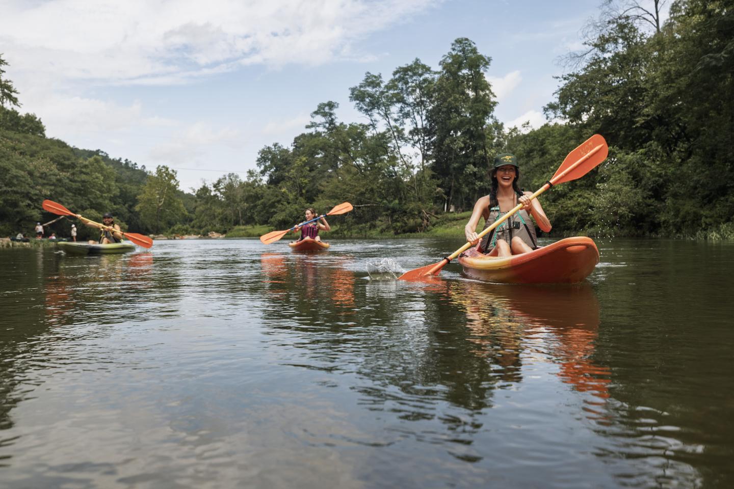 Three people kayaking on a calm river under a partly cloudy sky.