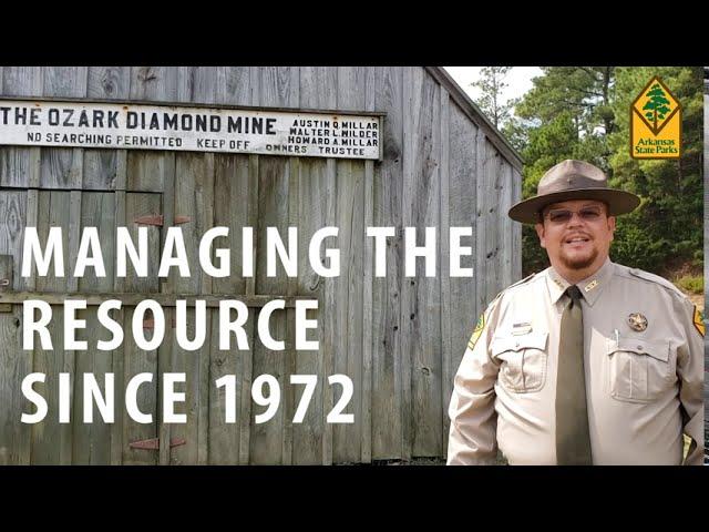 Park ranger stands beside a wooden cabin with a sign overhead.