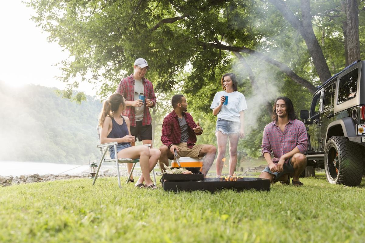 Group enjoying a barbecue by a river, surrounded by trees and a parked jeep.
