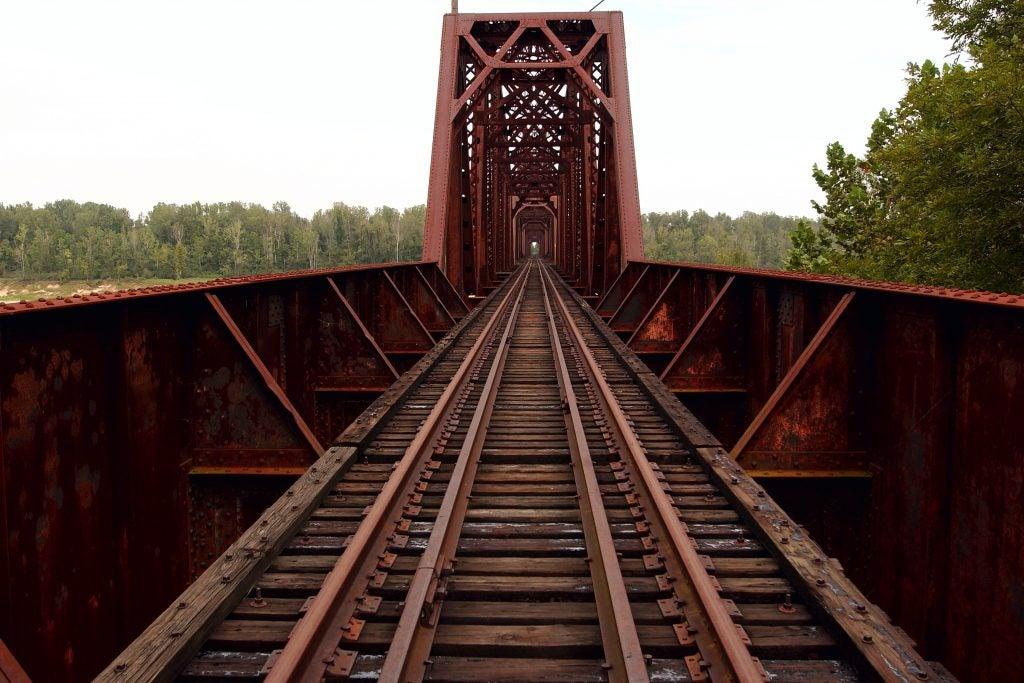 Railway tracks leading through a rusty metal bridge, surrounded by trees.