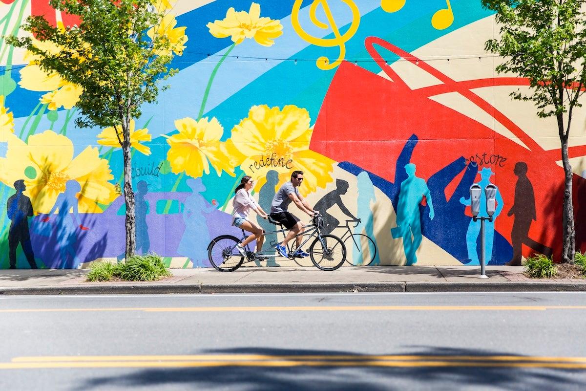 Two cyclists ride past a colorful mural with flowers and musical notes.