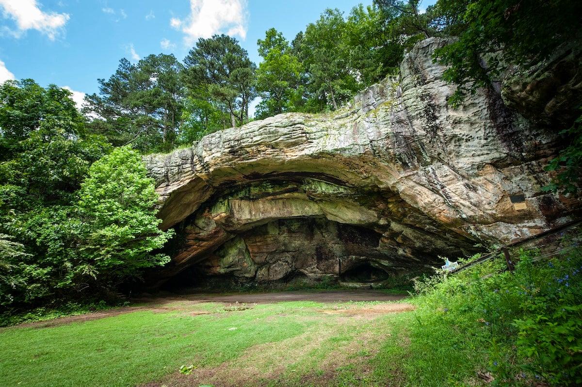 Rocky cave entrance surrounded by lush green trees under a blue sky.