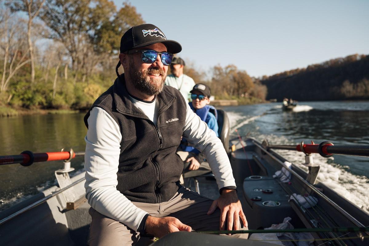 Man wearing sunglasses and cap, steering a boat on a sunny river.