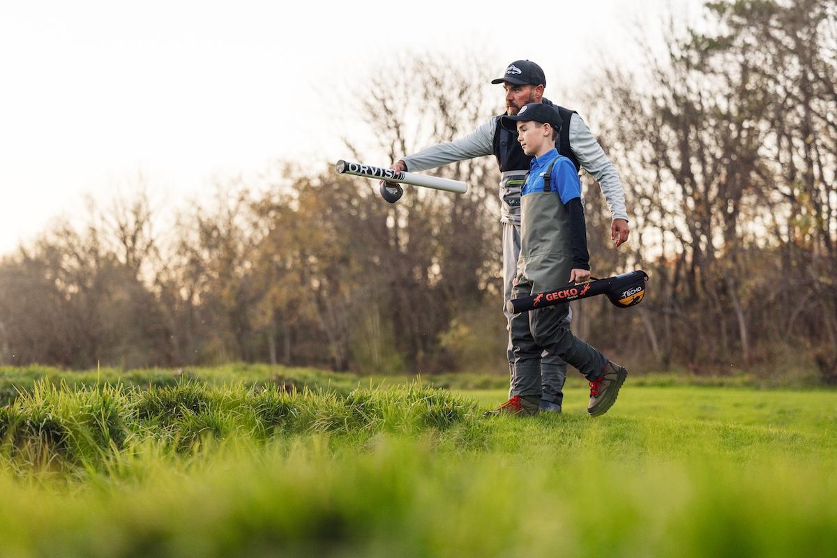 Father and son walking on grass, carrying fishing rods under a clear sky.
