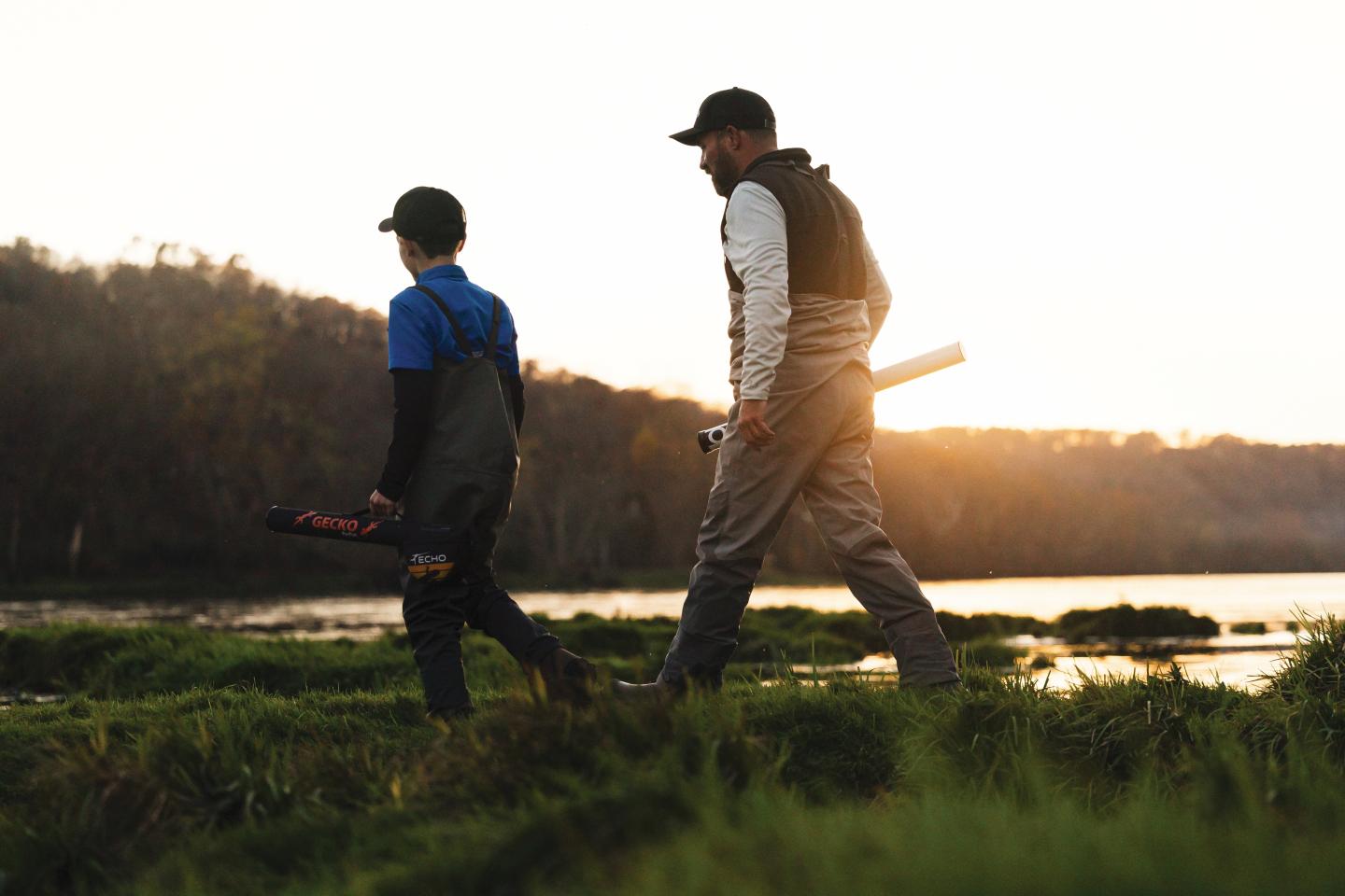 Two people walking on a grassy field at sunset, carrying fishing gear.