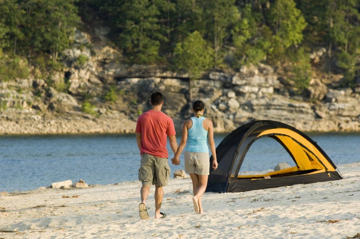 Couple walking on a sandy beach near a tent by a lake.