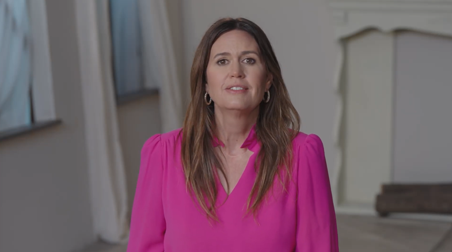 Woman in a bright pink shirt stands in a softly lit room with draped curtains.