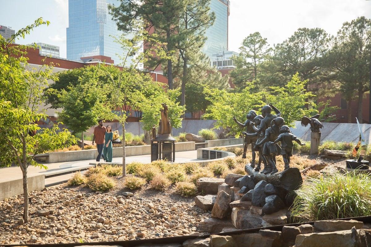 Sunny park with sculptures, trees, and city buildings in the background.