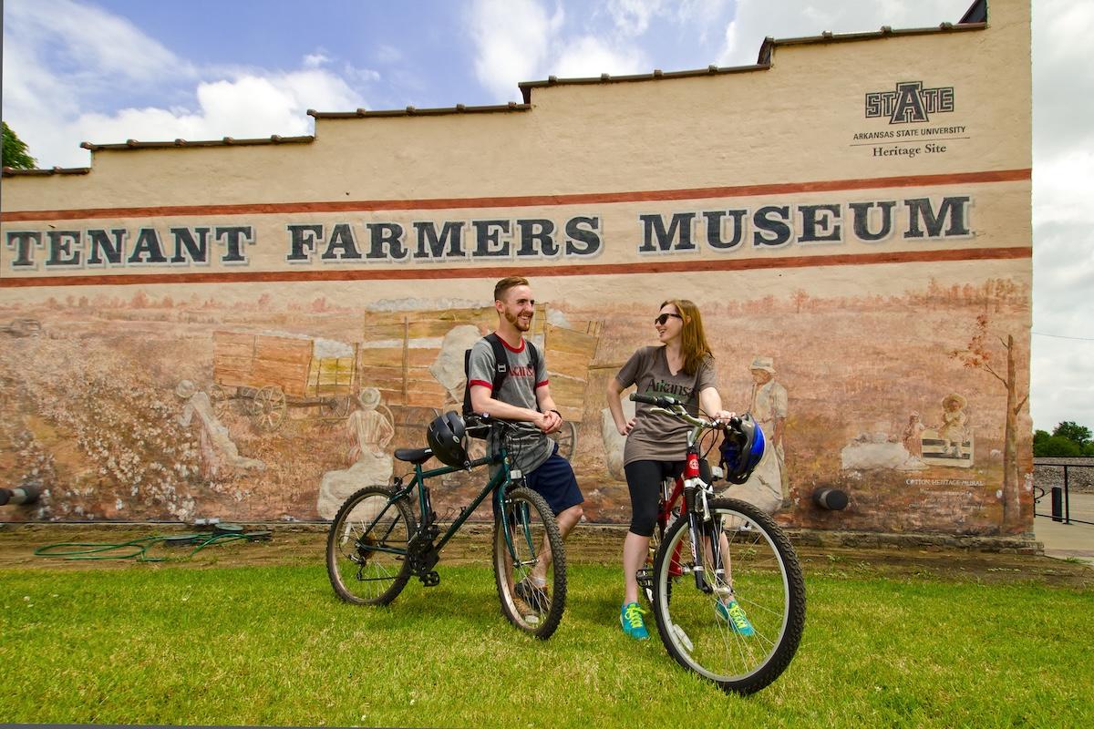 Two people with bicycles smiling in front of Tenant Farmers Museum wall.