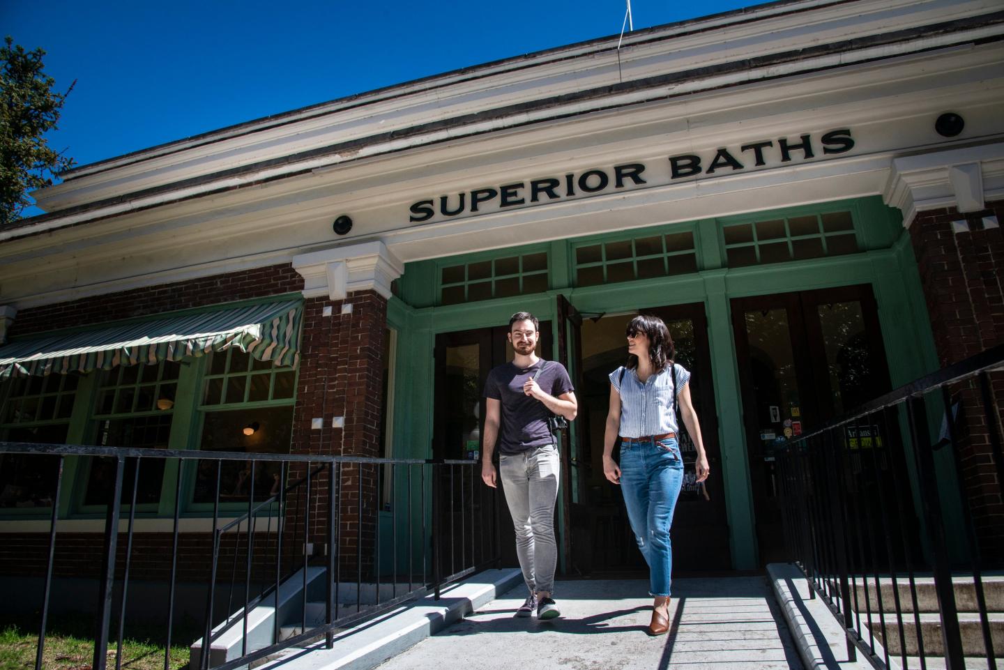 Two people walking out of a building labeled "Superior Baths" on a sunny day.