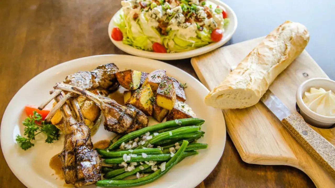 Lamb chops with green beans and potatoes, salad, and baguette on a wooden table.