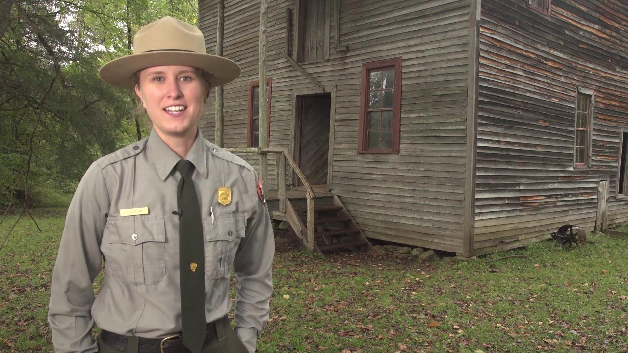 Park ranger smiling in front of a weathered wooden building outdoors.