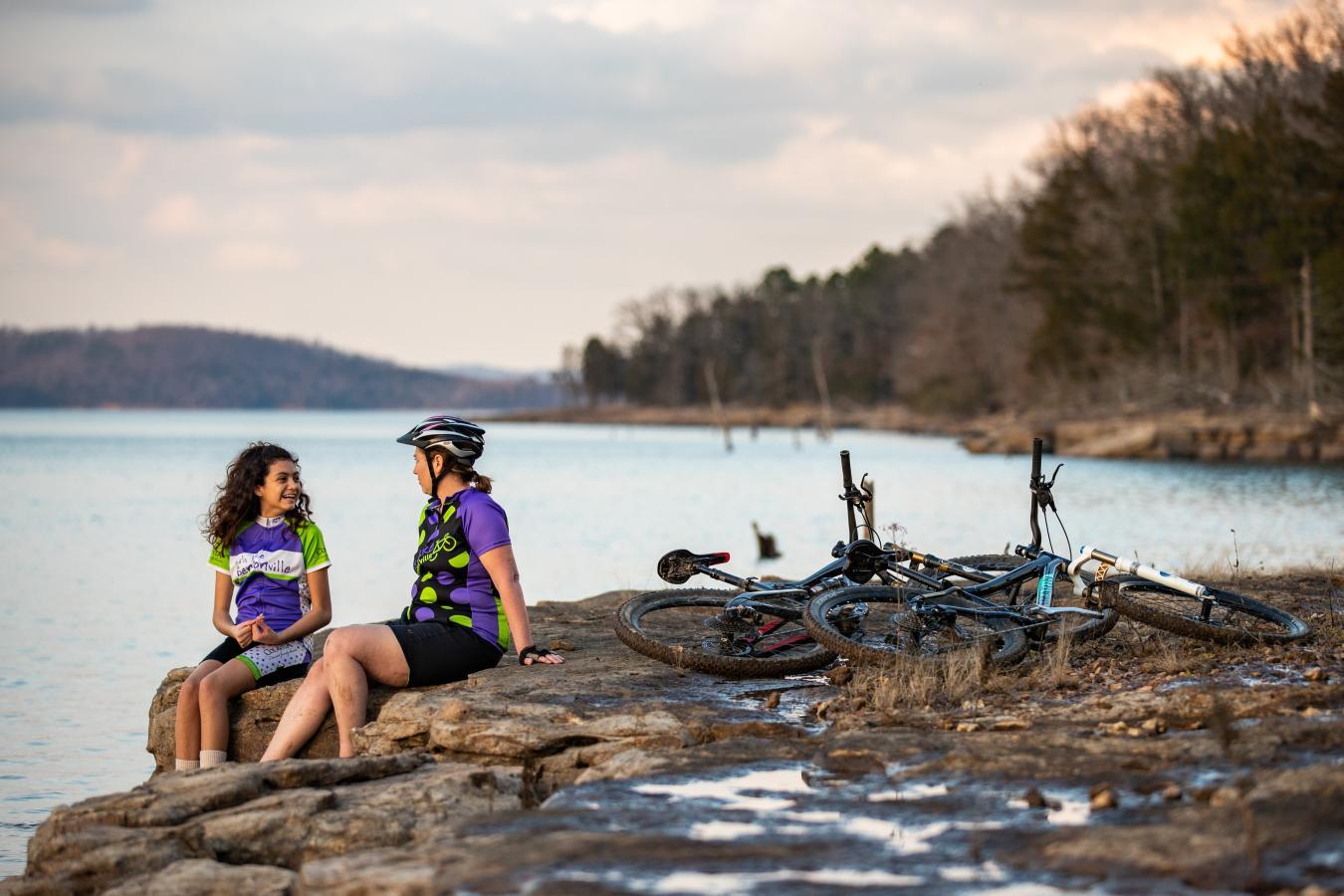 Two cyclists resting by a calm lake, bikes nearby, during sunset.