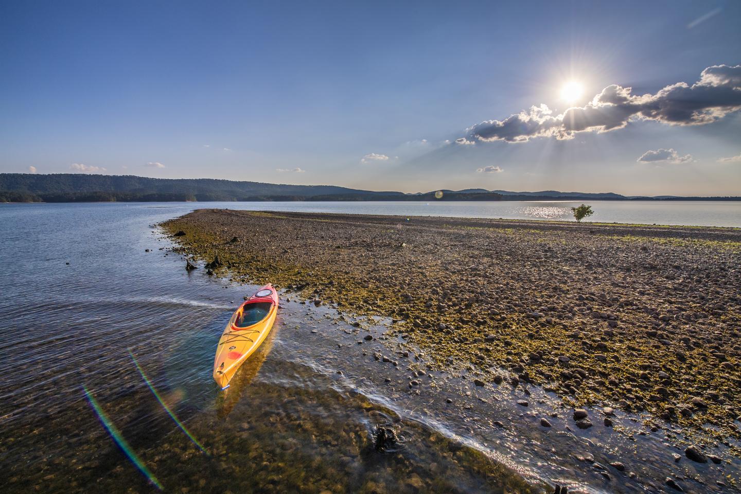 Yellow kayak on a calm lake under a bright sun, with pebbled shore nearby.