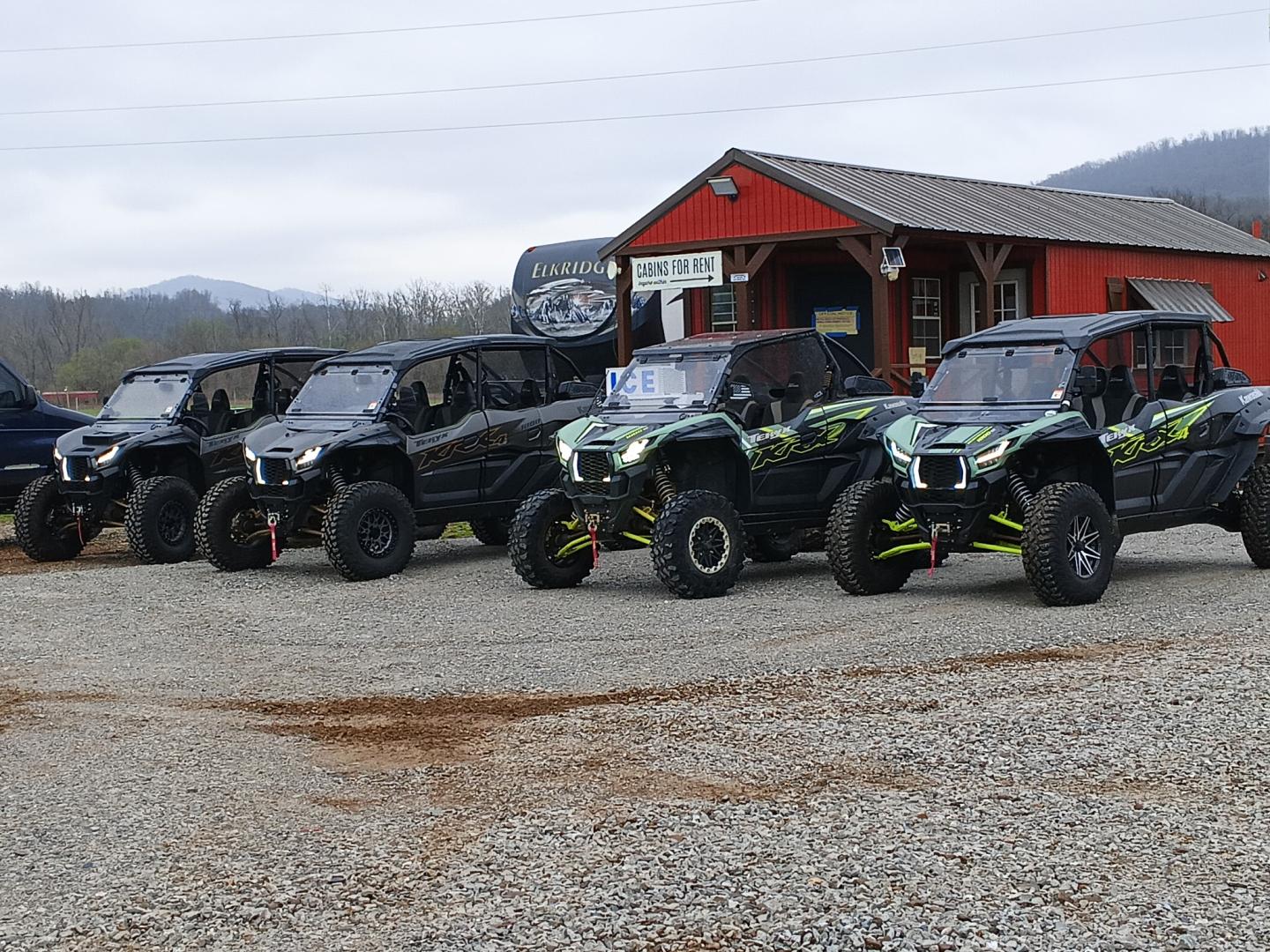 Five off-road vehicles parked near a small red building on gravel.