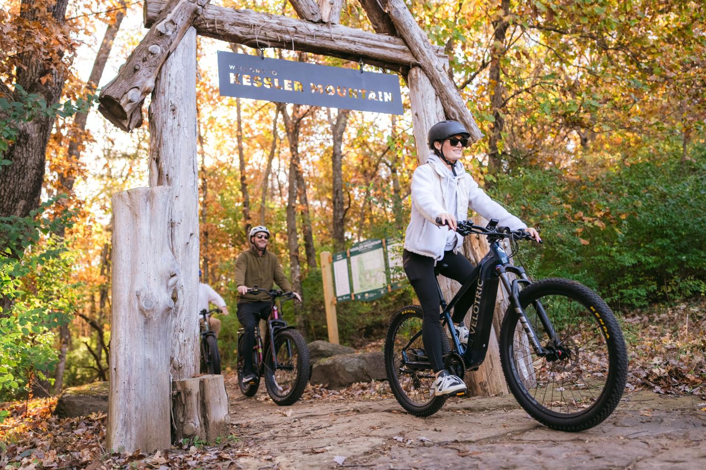 Cyclists in helmets riding through a forest archway in autumn.