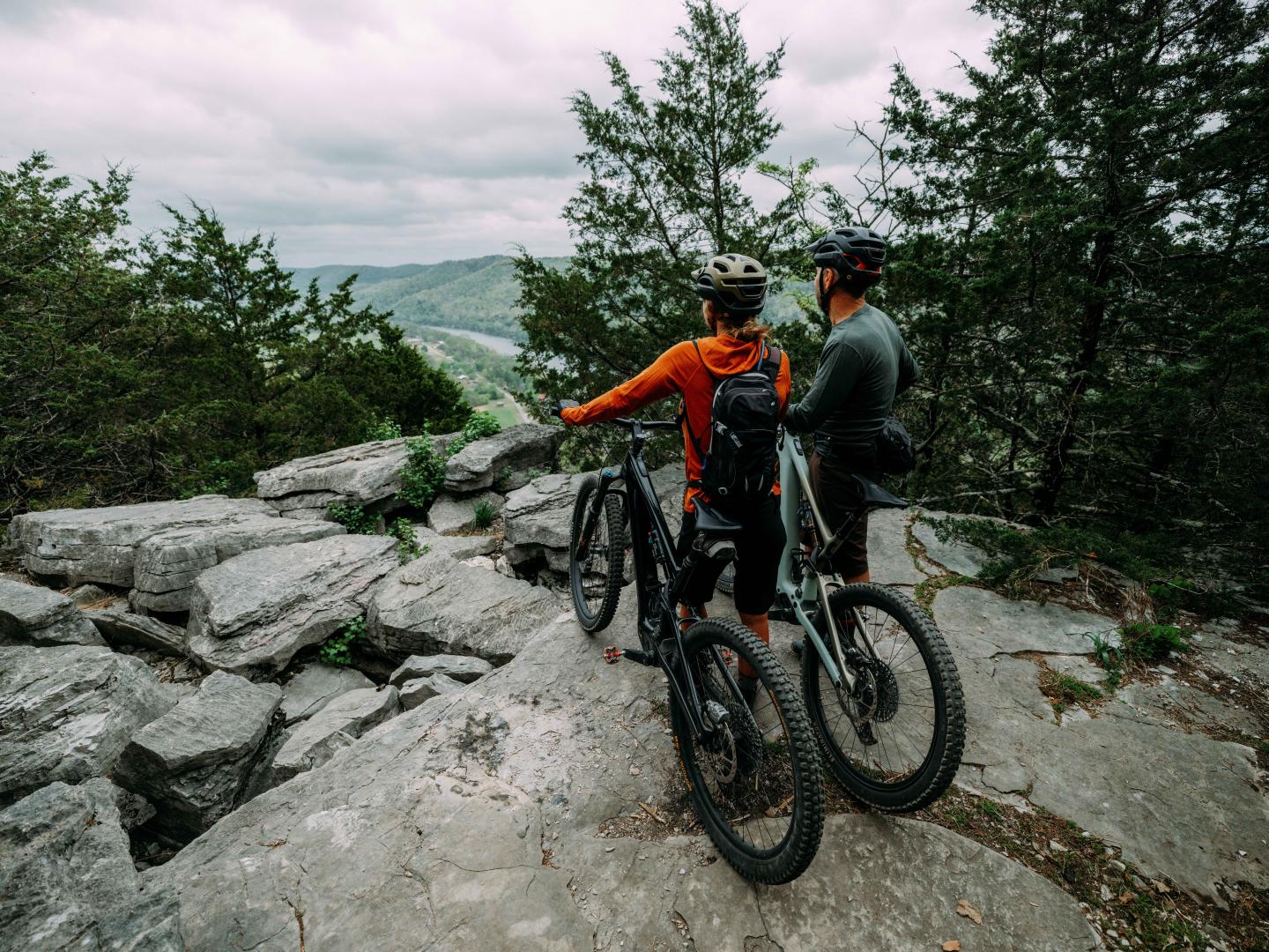 Two cyclists pause on a rocky trail surrounded by trees, overlooking a scenic valley.