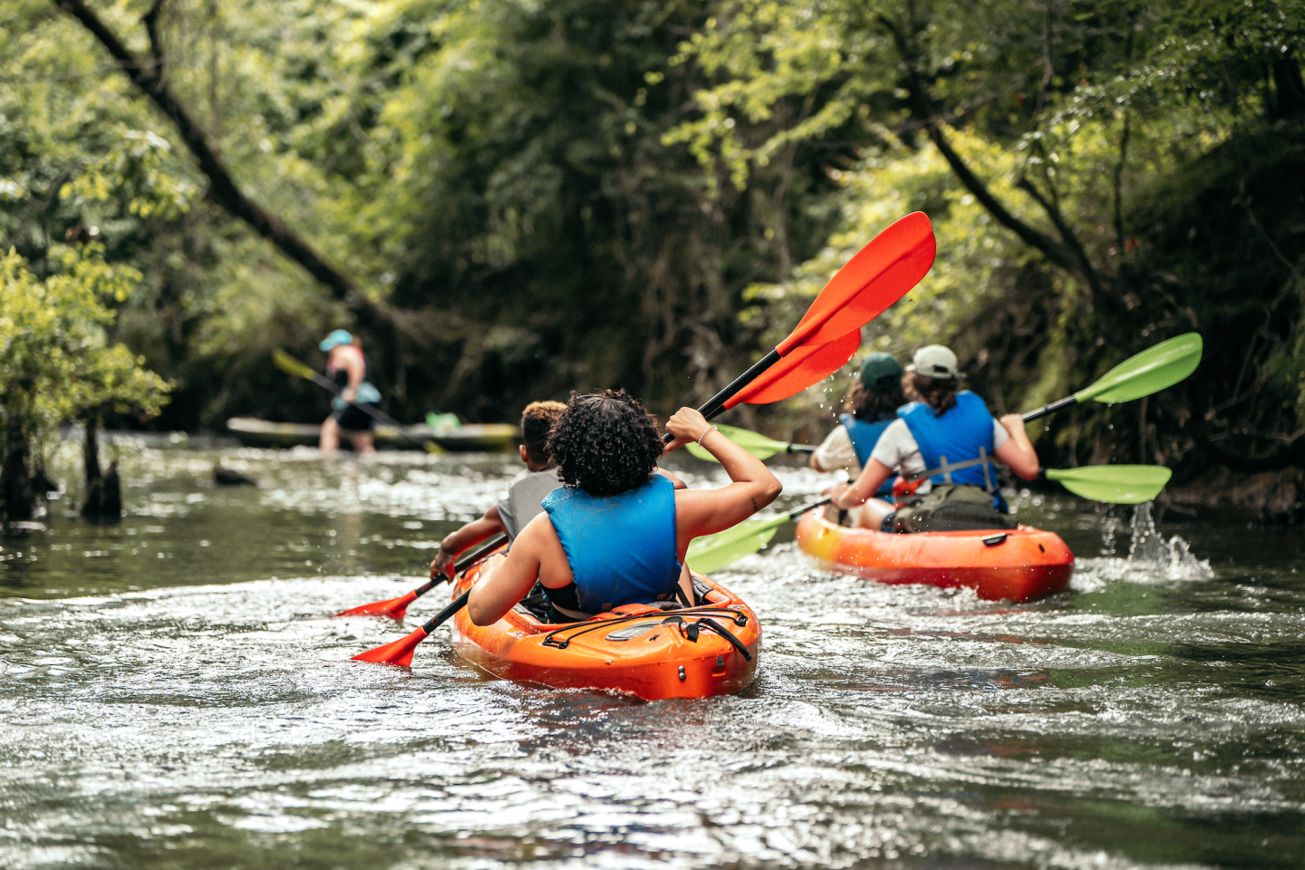 Kayakers paddle down a lush, tree-lined river.