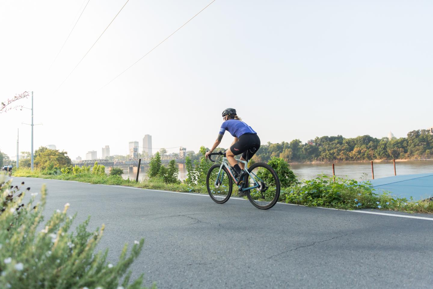Cyclist rides alongside a river with a city skyline in the distance.