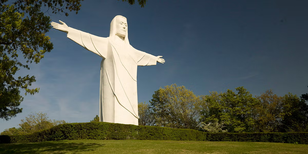 White statue of Jesus with outstretched arms against a blue sky, surrounded by trees.