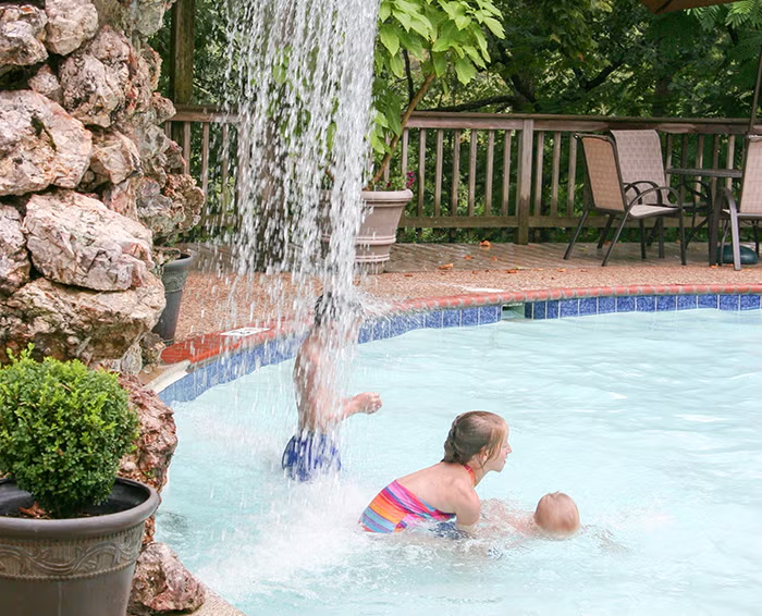 Children play beneath a waterfall in a swimming pool.