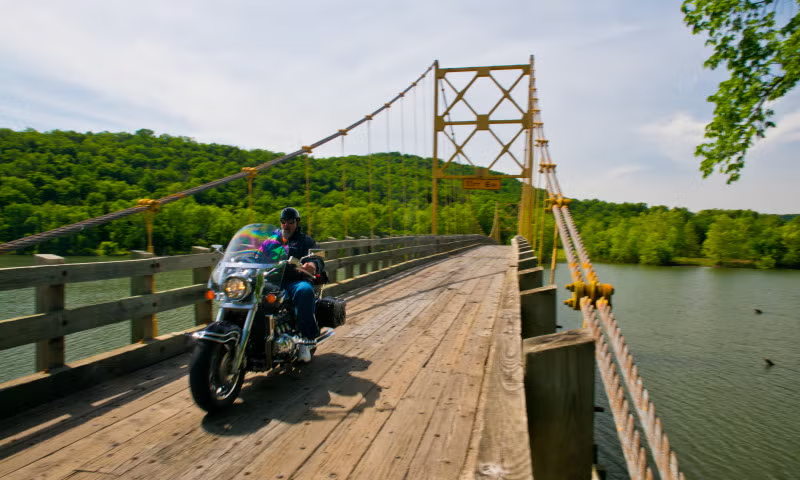 Motorcyclist crossing wooden bridge over a river, with green hills in the background.