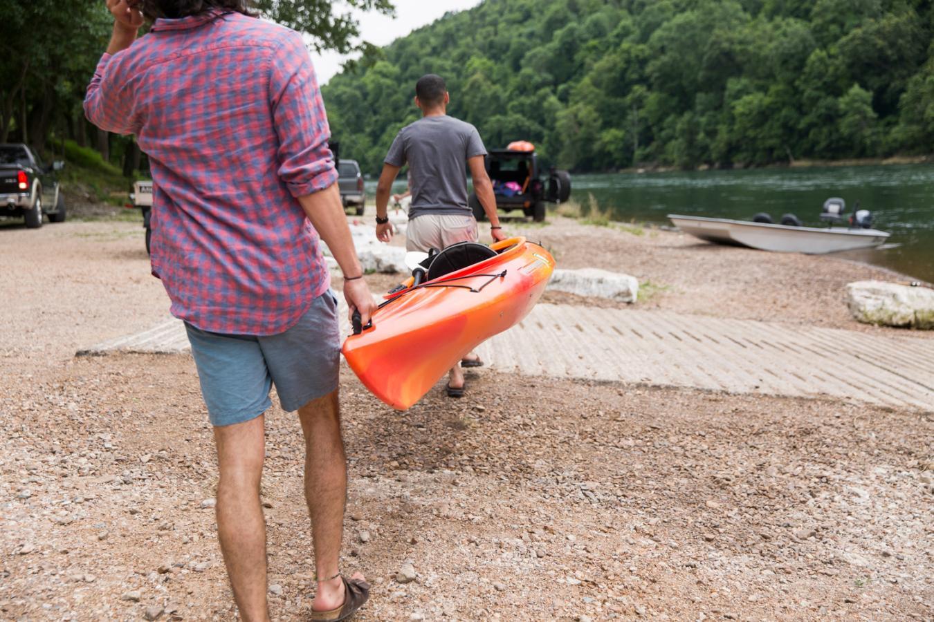 Men carrying a kayak to a riverside, surrounded by trees.