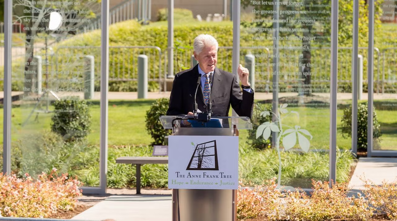 Man speaking at a podium outside with greenery in the background.