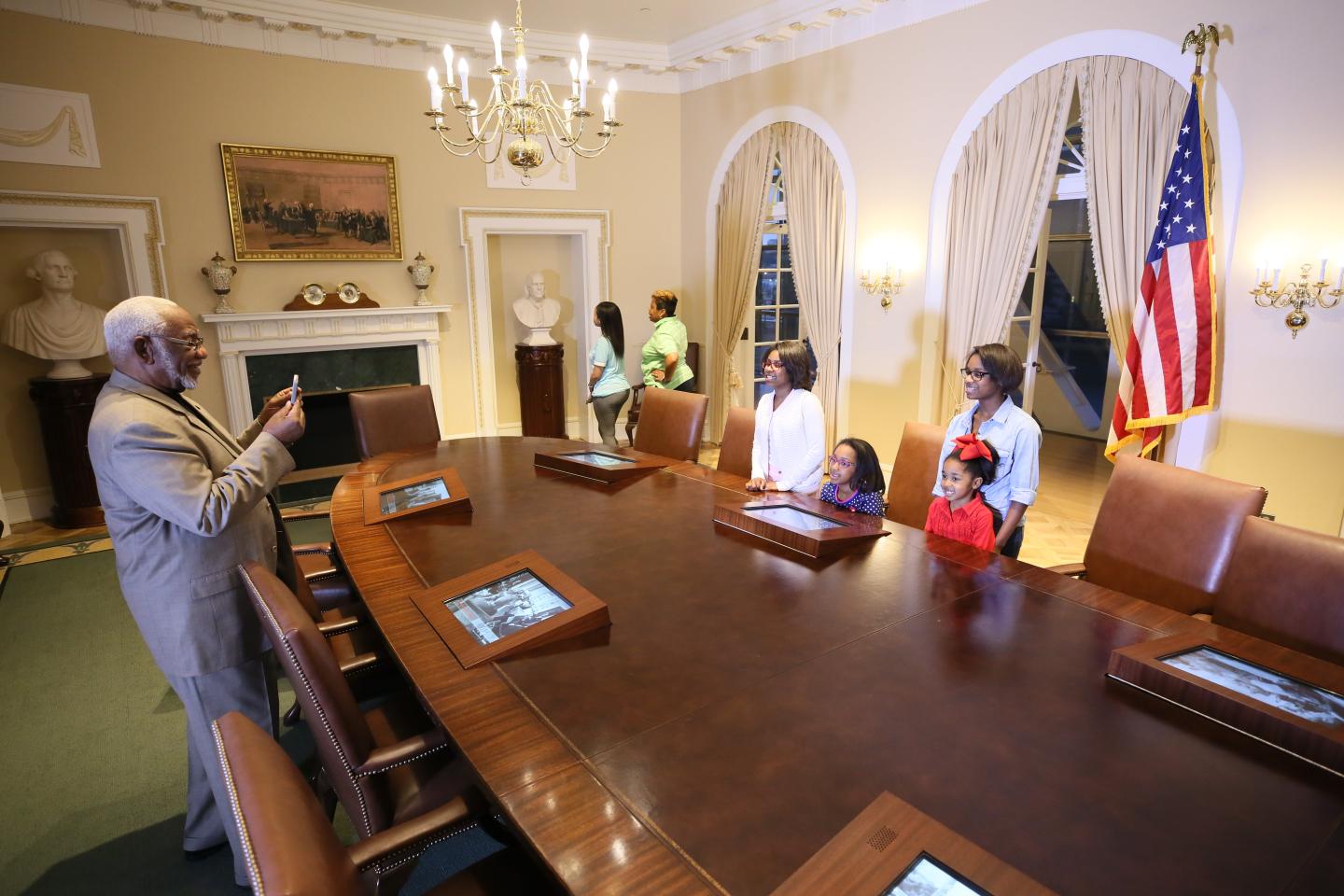 A group tours a historic room with a large wooden table and an American flag.
