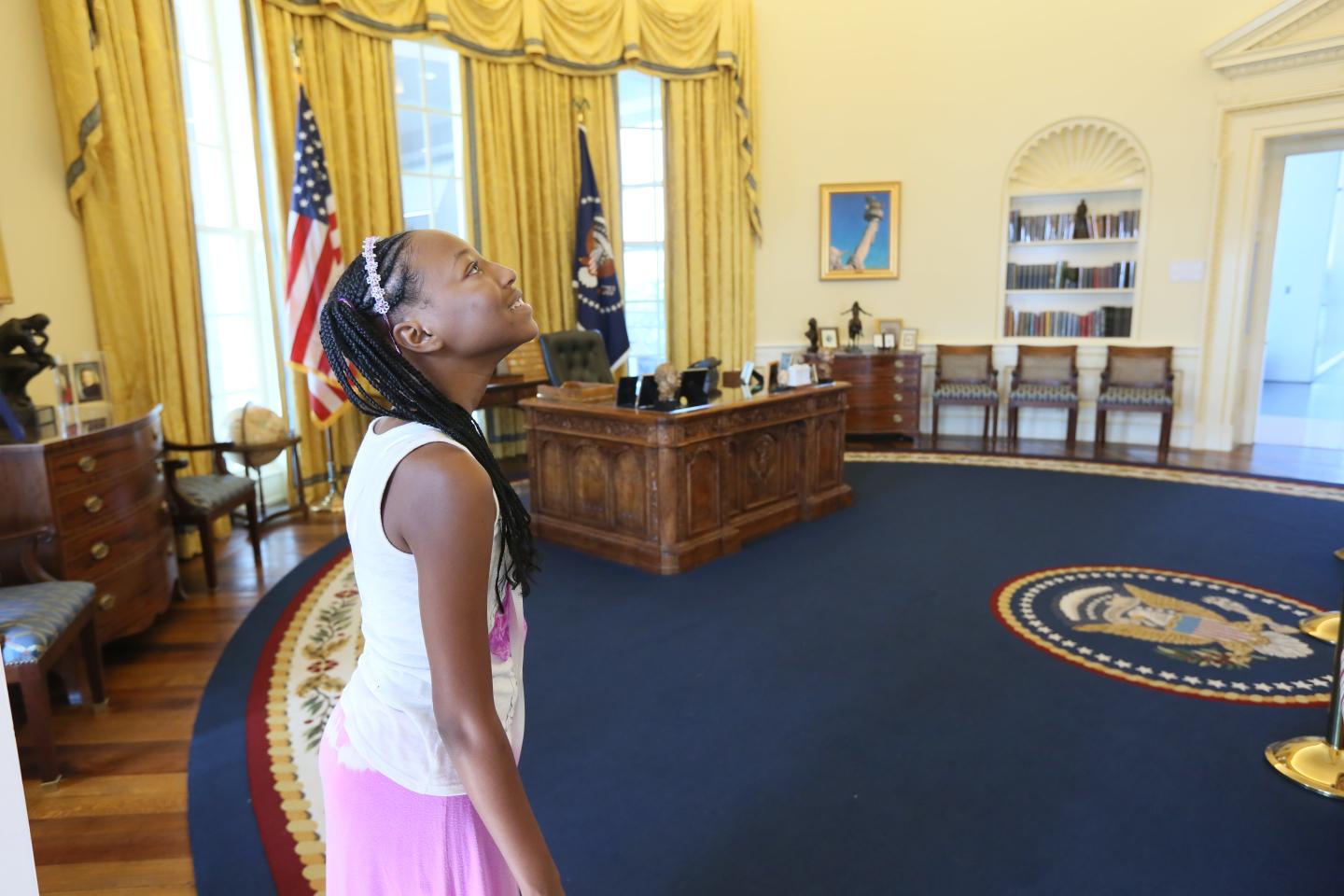 Girl in pink dress looking up in an ornate office with flags and a large desk.