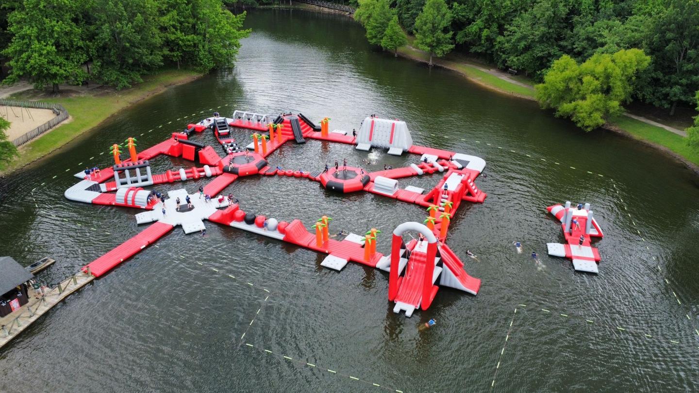 Floating red and gray inflatable water park on a lake, surrounded by trees.