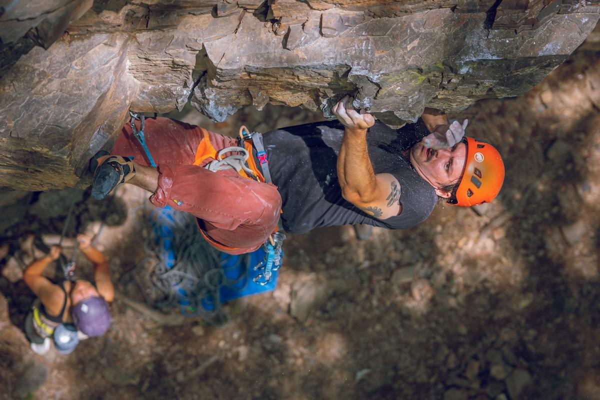 Rock climber in orange helmet ascends a wall, with another climber below on a sunny day.