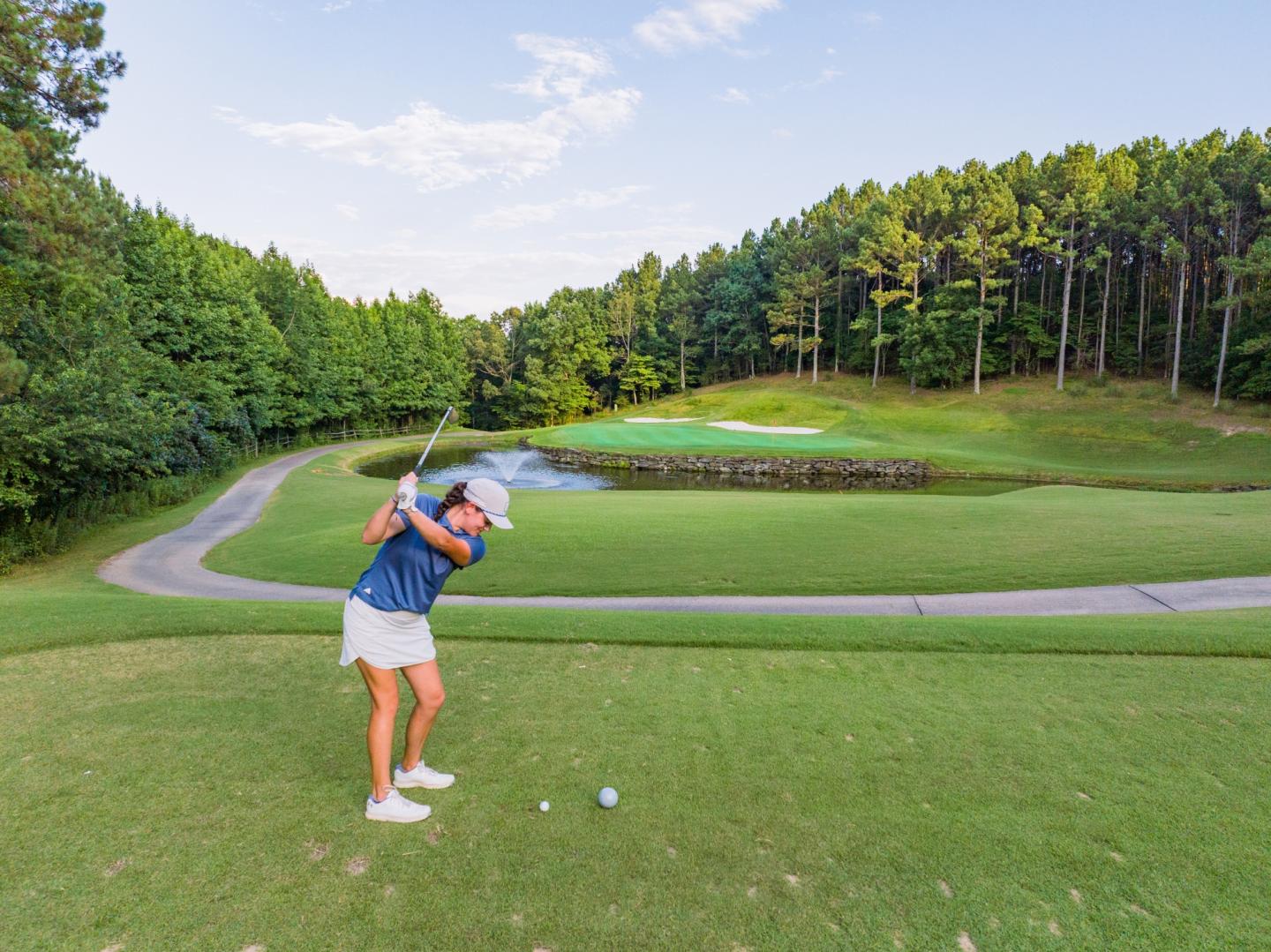 Golfer swinging on a lush, sunny course with trees in the background.