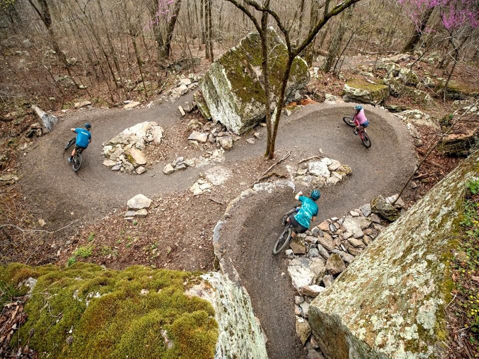 Three cyclists navigate a winding forest trail with large rocks.