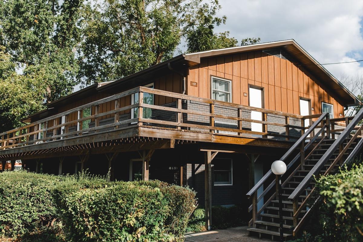 Two-story wooden cabin with a wraparound deck and green bushes.