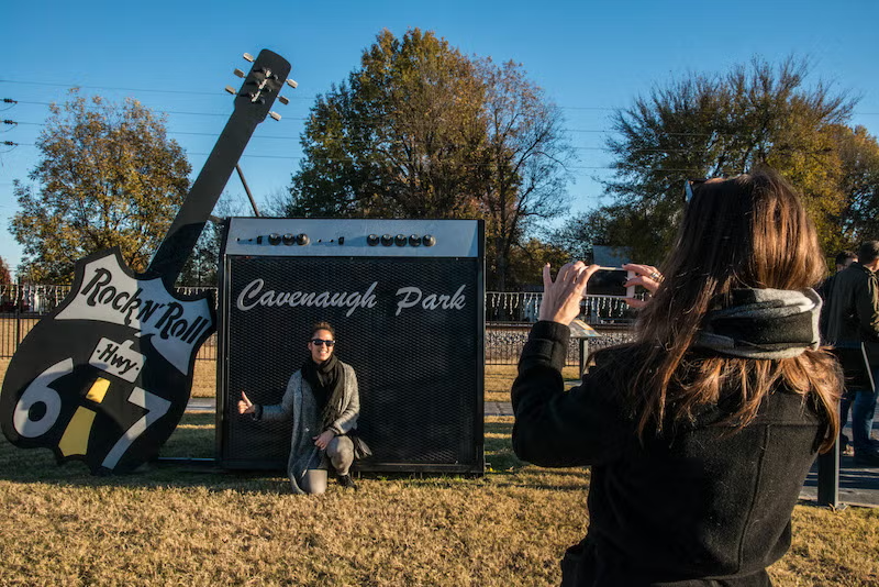 Large guitar sculpture, person kneeling, another taking photo, trees in background.