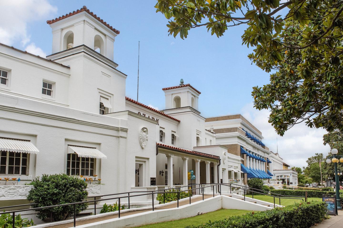 Elegant white building with columns, lamps, and nearby trees, under a partly cloudy sky.