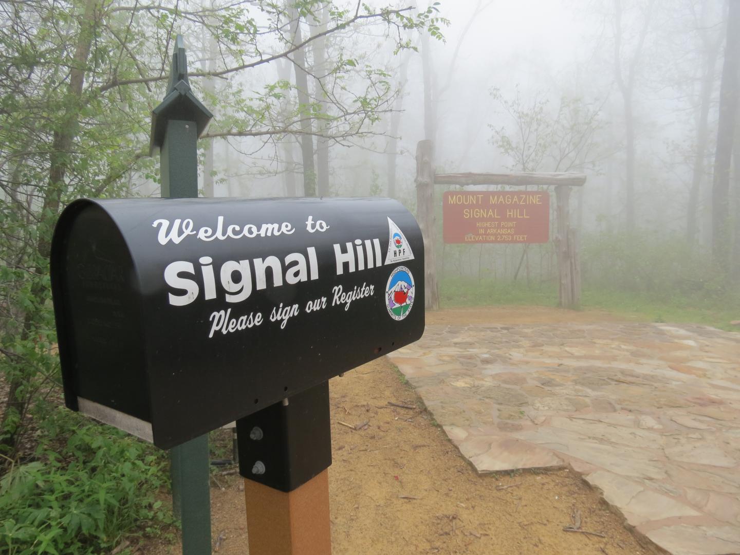 Mailbox by foggy forest path with park sign in background.