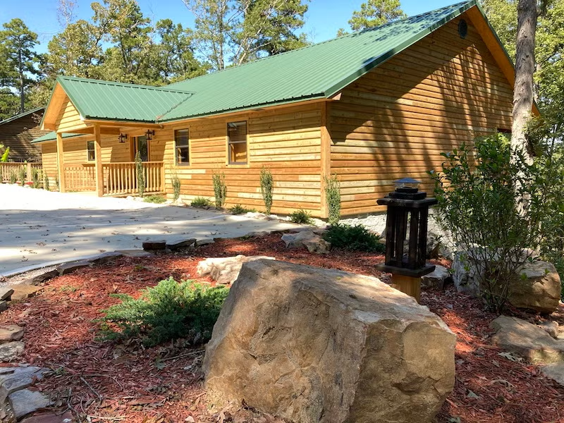 Cozy wooden cabin with green roof, surrounded by trees and stone landscaping.