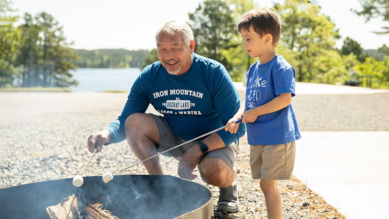Grandfather and boy roasting marshmallows by a lakeside campfire.