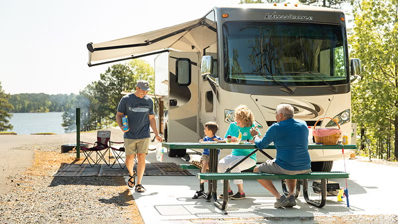 Family sitting at a picnic table near an RV by a lake, enjoying a sunny day.